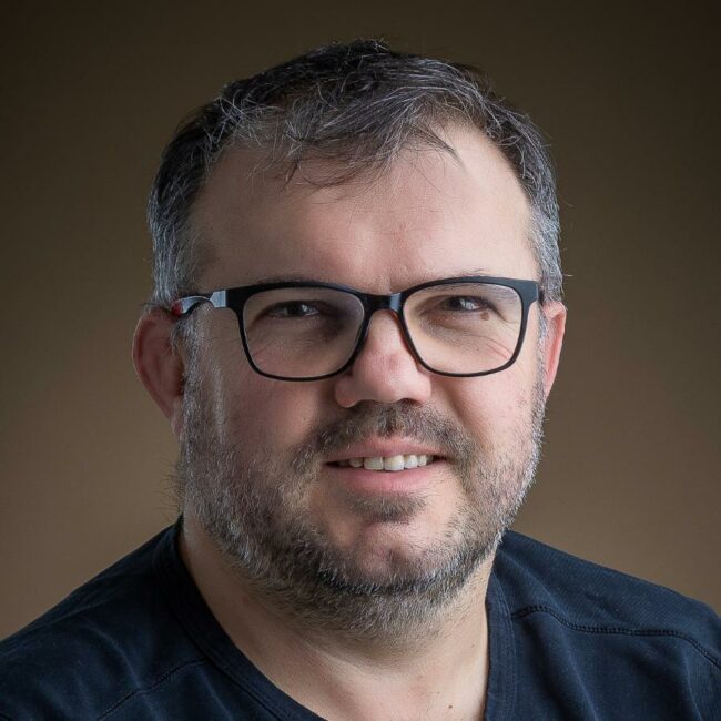 Portrait d'un homme avec des lunettes noires, une courte barbe et souriant.
